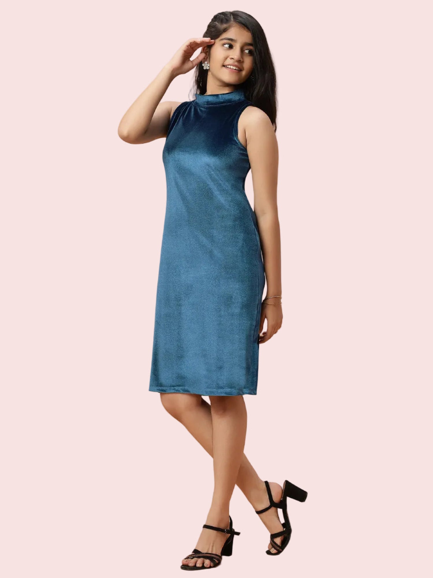 A woman modeling a sleeveless, high-neck blue dress with a velvet-like texture, paired with black strappy heels, set against a solid pale pink background.